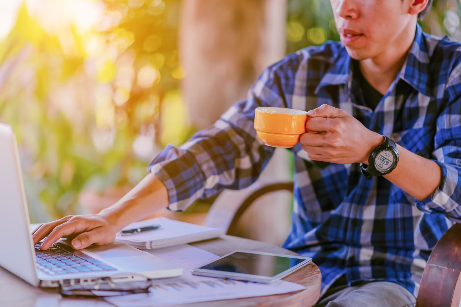A freelancer in a blue checkered shirt working on a laptop at an outdoor table in Switzerland, holding an orange coffee cup next to a tablet and financial charts, symbolizing a successful self-employment setup in 2026